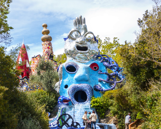 A giant blue surreal sculpture with an open mouth and a round red eye, part of an outdoor park installation; a silver top piece and other whimsical structures rise behind it, with trees and a few visitors standing nearby.