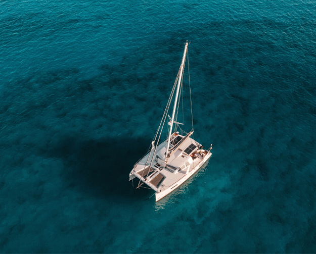 An aerial view of a white sailboat with a tall mast on calm turquoise sea.