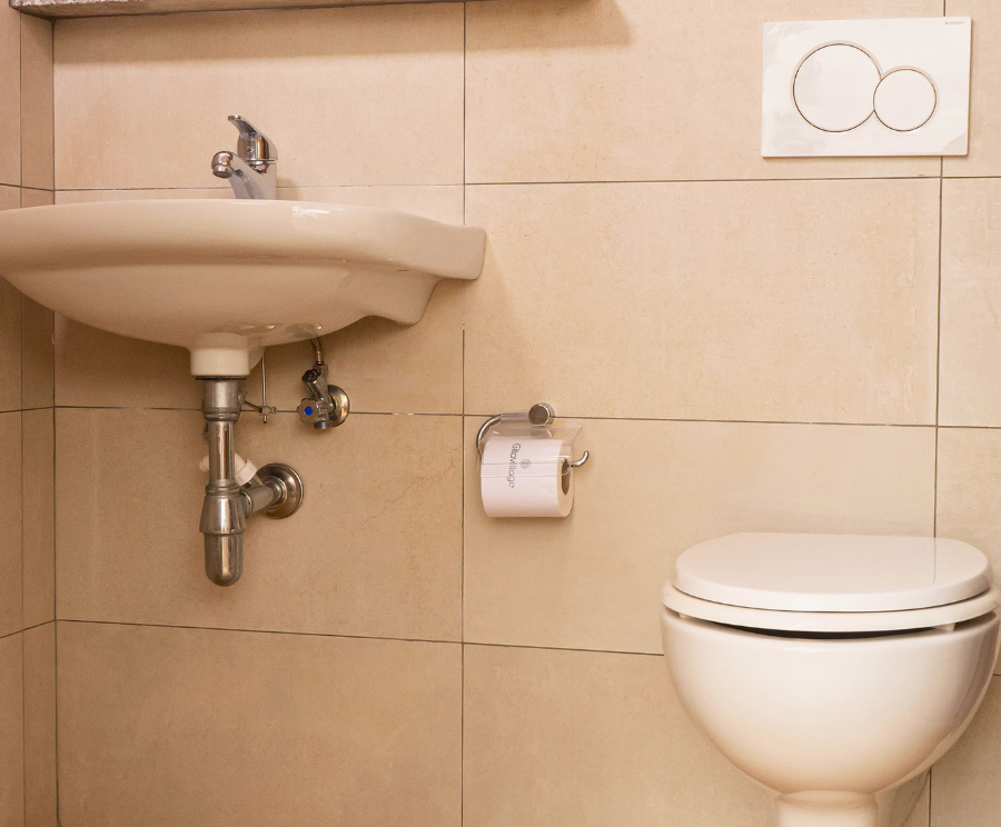 Bagno con lavabo a sinistra e WC a destra; sul muro, pulsante di scarico a due tasti e porta carta igienica; pareti piastrellate beige.