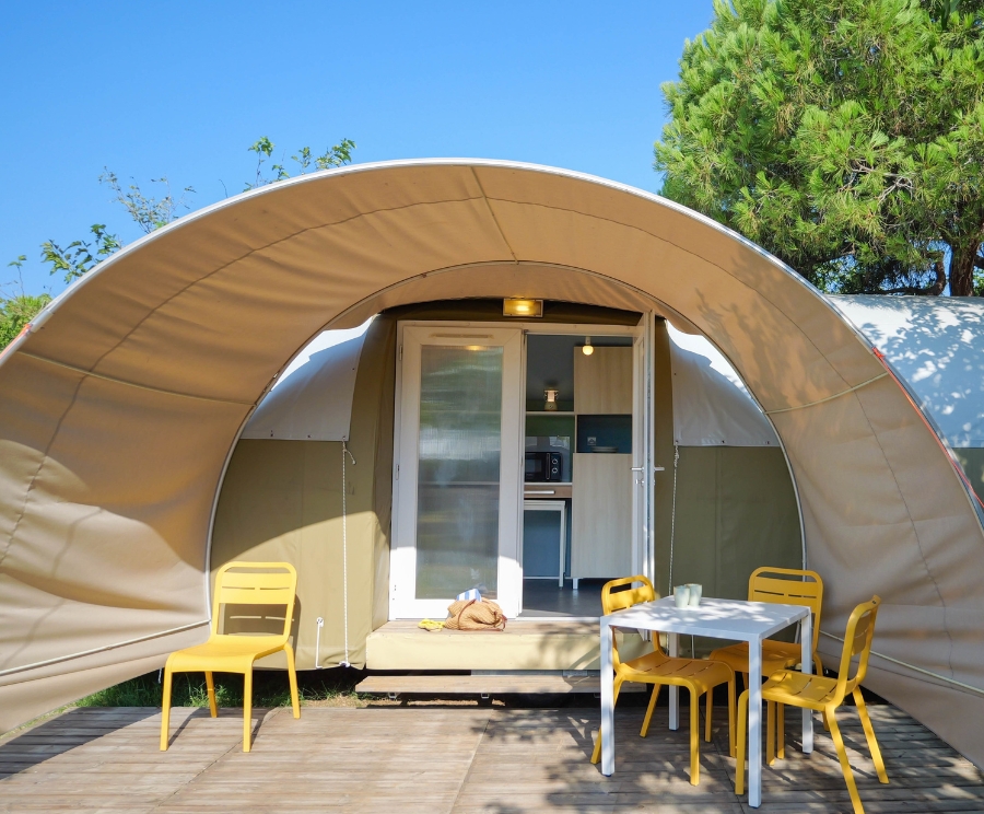 Una grande tenda a cupola beige con ingresso in vetro si apre su una terrazza di legno. Davanti all'ingresso ci sono un tavolo bianco e sedie gialle, e una cagnolina è distesa sui gradini. Sullo sfondo, cielo azzurro e alberi verdi.