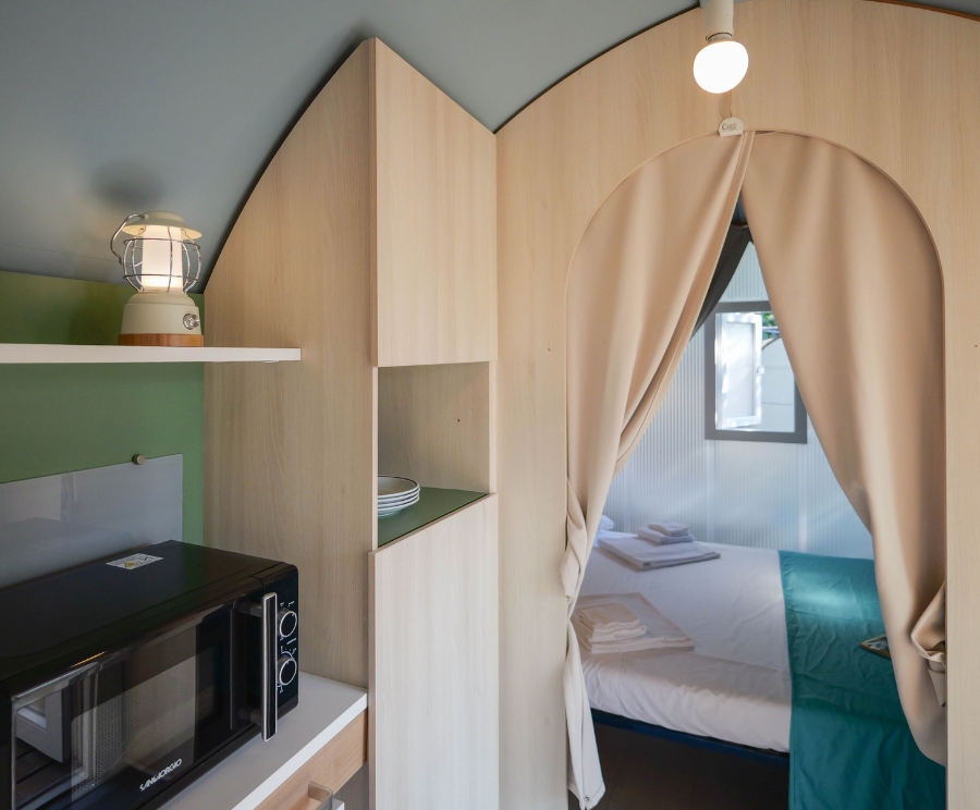 Kleines Zimmer mit Holzmöbeln: links eine Mikrowelle unter einem Regal, daneben ein offenes Holzregal und eine Laterne. Durch einen halbrunden Vorhangbogen führt der Durchgang ins Schlafzimmer; dahinter befindet sich ein Bett mit weißer Bettwäsche und türkisfarbenem Überwurf. Ein Fenster ist hinter dem Vorhang zu sehen.