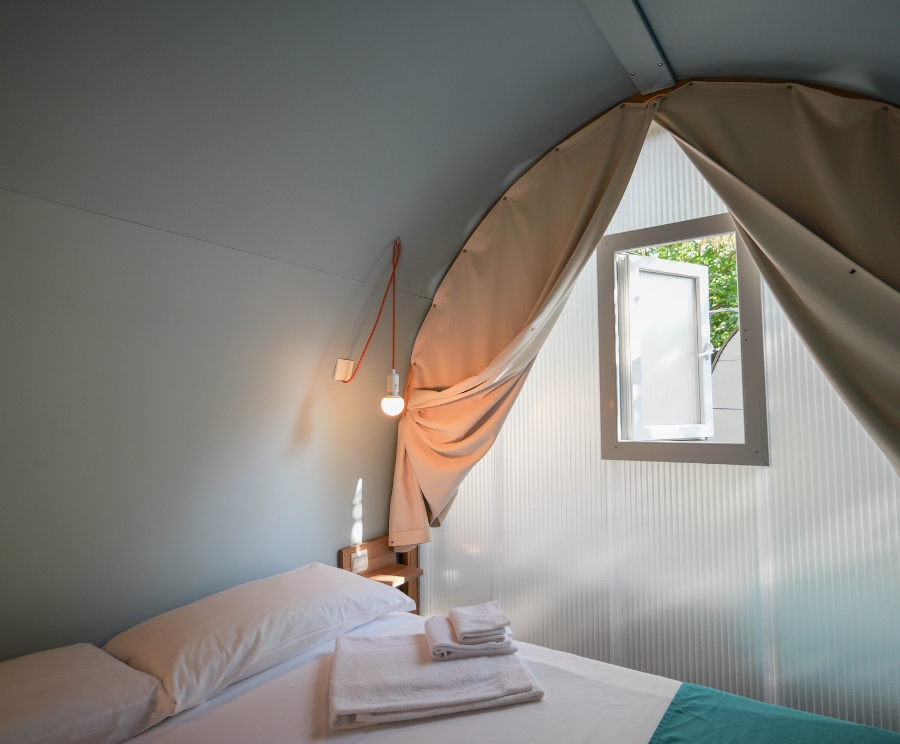 Interno di una tenda trasformata in una camera: letto matrimoniale con lenzuola bianche e asciugamani piegati, una lampadina sospesa, un comodino in legno e una finestra aperta che lascia entrare la luce esterna.