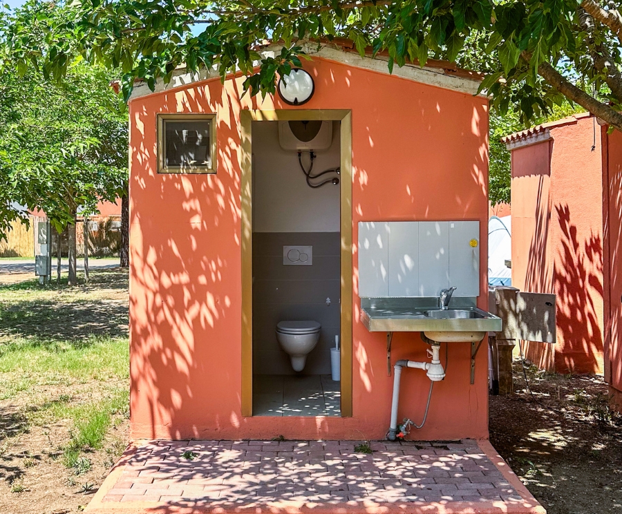 Piccolo bagno pubblico arancione con porta aperta: si intravede una toilette all'interno; accanto c'è un lavabo esterno con tubi visibili. L'area è ombreggiata dagli alberi e pavimentata con mattoni.