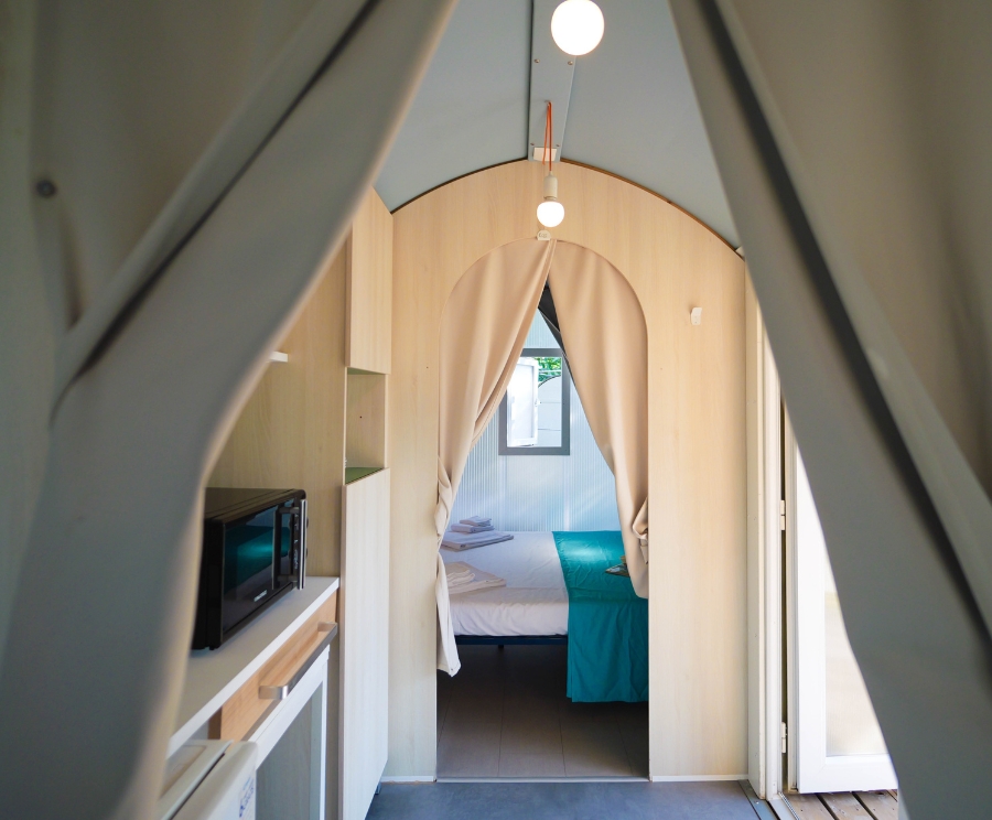 Interno di una cabina: cucina piccola a sinistra con microonde e scaffali, un corridoio con tende che formano un arco conduce a una camera da letto sullo sfondo con letto bianco e coperta azzurra.
