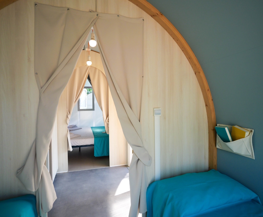 Interno di una camera a forma di yurta con arco in legno e tende bianche: corridoio che conduce a una seconda stanza, letto con copriletto azzurro sulla destra, e una nicchia porta-oggetti fissata alla parete; una luce pendente illumina l'ambiente.