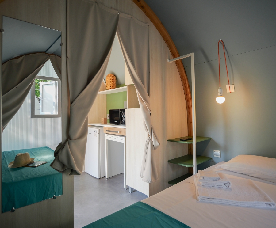 Interno di una piccola cabina: a destra un letto con lenzuola bianche, a sinistra un secondo letto coperto da una coperta verde acqua. Una tenda separa la zona notte dalla cucina, dotata di frigorifero, microonde e ripiani verdi. Una lampadina esposta pende dal soffitto con un filo rosso. Un arco di legno delimita lo spazio e un cappello è posato sul letto di sinistra.