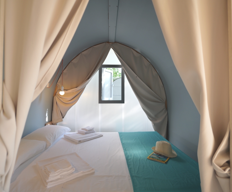 Camera da letto all'interno di una tenda a teepee: letto matrimoniale con lenzuola bianche, coperta azzurra sul lato destro, asciugamani piegati sul letto, cappello e riviste sulla coperta; lampada appesa sul lato sinistro e una finestra rettangolare al centro della parete posteriore, tende beige che incorniciano lo spazio.