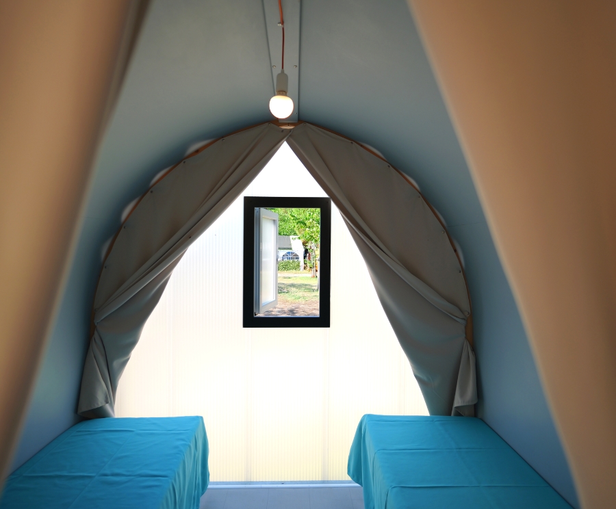 Interno di una tenda a forma di arco con pareti azzurre, due panche coperte di blu ai lati, una piccola finestra rettangolare al centro sullo sfondo e una lampadina appesa al soffitto.