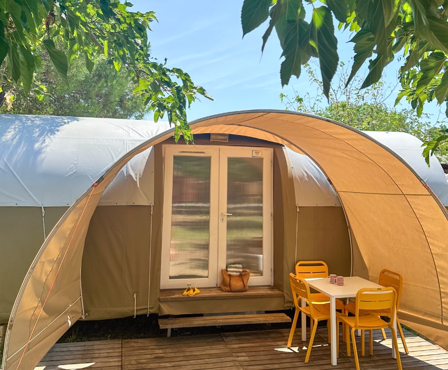 Una tenda glamping a cupola beige con porta in vetro è montata su una pedana in legno. Accanto, tavolo bianco con sedie gialle, e sullo sfondo si vede un giardino soleggiato con foglie verdi e cielo azzurro.