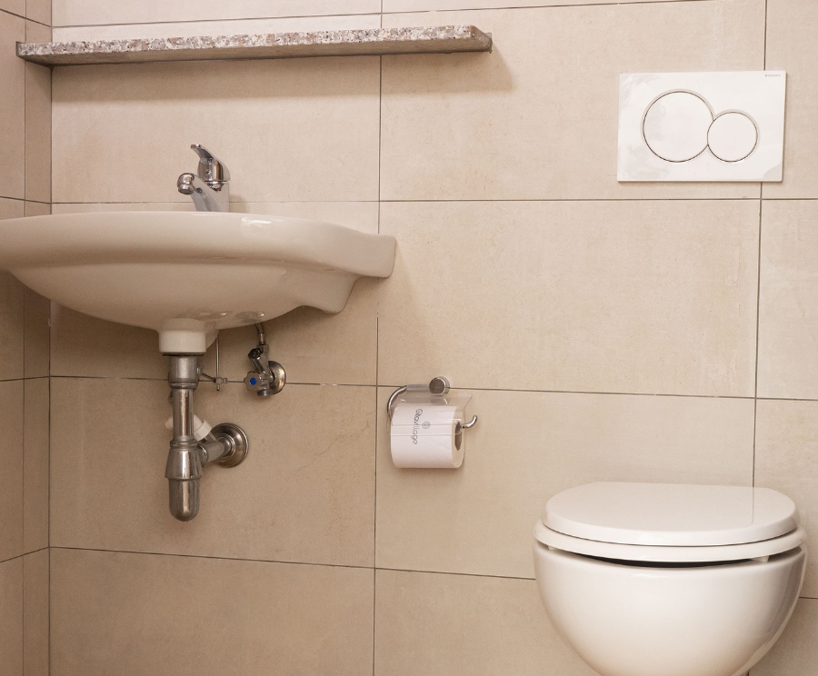 Ein Badezimmer mit einem wandhängenden Waschbecken, darunter sichtbare Rohre, einer Toilette, einem Toilettenpapierhalter und einem Spülkasten an der Wand mit zwei runden Knöpfen; die Wände sind hellbeige gefliest.