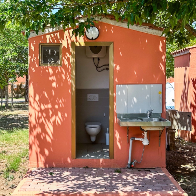 Cabina igienica esterna color pesca con la porta aperta che mostra un water all'interno; a destra è fissato un lavabo in acciaio con tubature visibili. Ombre di foglie sulle pareti e sul pavimento in mattoni, in un cortile soleggiato.
