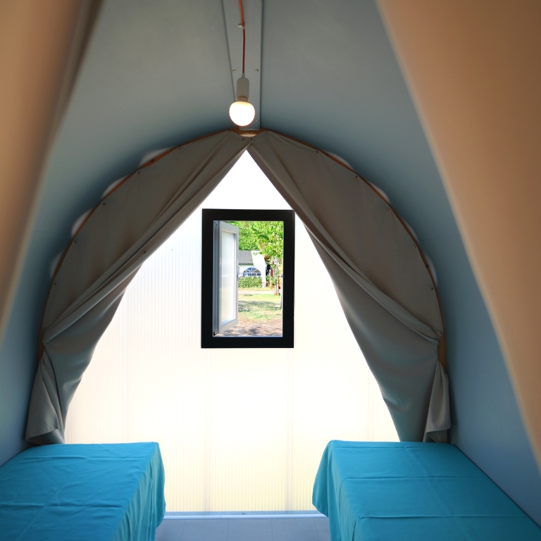 Interno di una tenda a forma di arco con pareti chiare; ai lati due tavoli rivestiti di tessuto azzurro, una piccola finestra nera al centro e una lampadina appesa al soffitto.