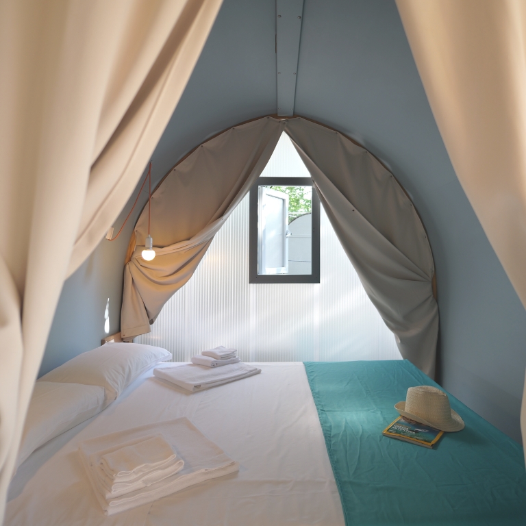 Una camera da letto in stile tendone con letto matrimoniale bianco, runner turchese, tende beige che incorniciano l’ingresso, una piccola finestra triangolare sullo sfondo, una lampadina sospesa a sinistra, asciugamani piegati sul letto e un cappello di paglia accanto a una rivista sul lato destro.